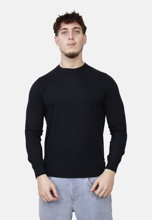 Giovane uomo con capelli corti e ricci, che indossa una maglia nera a maniche lunghe e jeans azzurri, in piedi davanti a uno sfondo bianco.