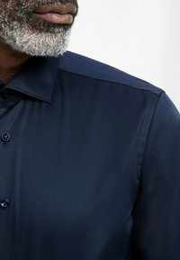 Camicia blu navy con una texture liscia, colletto con bottoni e un singolo bottone visibile sullo sbieco; design minimalista senza fantasie.