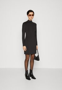 Robe longue noire à manches longues avec un col haut, une silhouette ajustée et une texture lisse. Associée à des bottines noires et un petit sac à main noir.