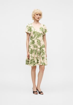 Femme aux cheveux blonds bouclés portant une robe beige mi-longue avec des imprimés de feuilles vertes et des chaussures à talons noires ouvertes à l'avant, debout sur un fond uni.