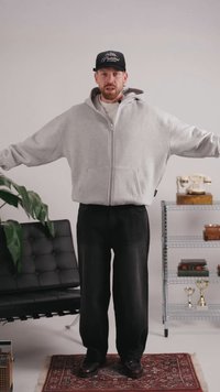 Homme en sweat à capuche gris et casquette noire debout, les bras étendus, dans une pièce avec une chaise noire, une plante, un téléphone vintage et une étagère métallique avec des trophées.