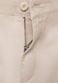 Pantalons kaki avec une fermeture éclair visible à dents métalliques, un bouton de fermeture beige et des détails de couture le long de la taille et de la zone de la fermeture éclair.