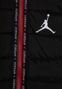 Czarna pikowana tkanina z czerwonym zamkiem błyskawicznym i czarnymi paskami, na których powtarza się tekst "Jordan" oraz logo Jumpman haftowane na biało.