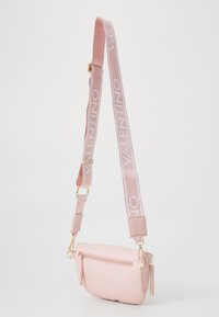 Rozā ādas plecu soma ar teksturētu virsmu, zelta aksesuāriem un platu siksnu, uz kuras baltā krāsā ir uzraksts "VALENTINO".