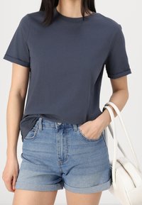Navyblauw katoen t-shirt met korte opgerolde mouwen, gecombineerd met lichtblauwe denim shorts. Het model houdt een witte handtas vast.