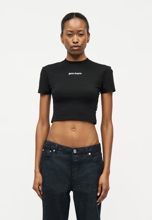 Γυναίκα που στέκεται μπροστά φορώντας μαύρο κοντό μπλουζάκι με το κείμενο «palm angels» και σκούρο μπλε ψηλόμεσο τζιν, με φόντο λευκό.