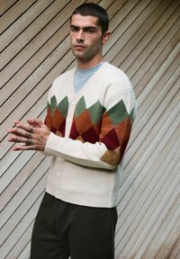 Kreemikardigan rohelise, oranži ja burgundia värvi argüülmustriga. Omab V-kaela lõike, nööbiga eest ja soonikkoes mansette.