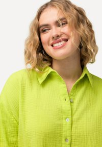 Camicia button-up verde lime con un tessuto testurizzato e stropicciato. Presenta un colletto e bottoni visibili, con un design casual.