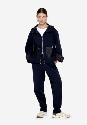Sudadera con cremallera de color navy con acentos de bolsillo de cuero y puños estampados, combinada con pantalones de chándal a juego y zapatillas ligeras.