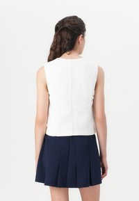 Witte mouwloze top met een strak ontwerp, voorzien van een rechte zoom en een naad aan de achterkant. Gecombineerd met een marineblauwe geplooide rok voor contrast.