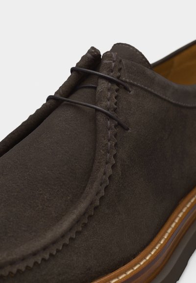 Σκούρο καφέ μοκασίνι από σουέντ με ραφές moc toe, λεπτή δερμάτινη κορδέλα και ταμπά σόλα με λευκή διακοσμητική ραφή.