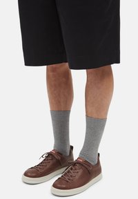Brown leather sneakers with a white sole, gray ribbed socks, and black knee-length shorts. Features include lacing and a red logo.