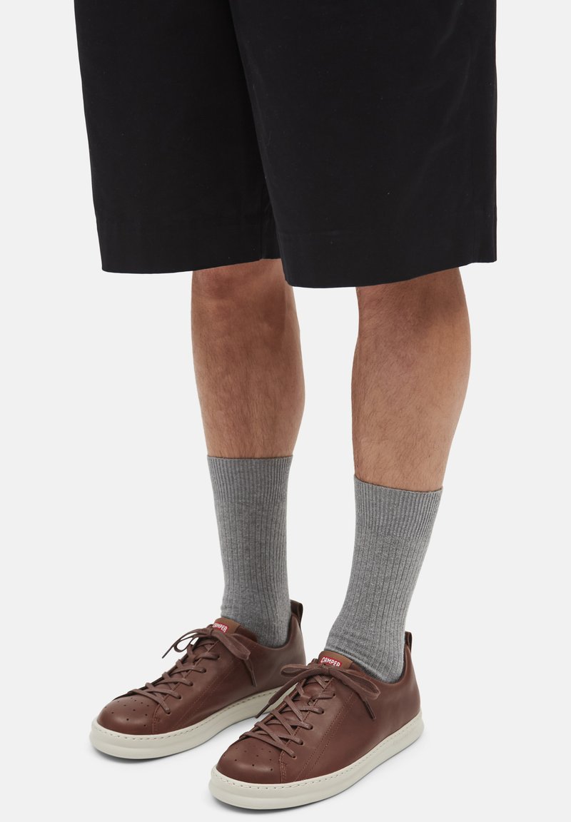 Brown leather sneakers with a white sole, gray ribbed socks, and black knee-length shorts. Features include lacing and a red logo.