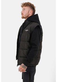 Zwarte quiltedvest met hoge kraag, ritsluiting en mouwloos ontwerp, gedragen over een zwarte hoodie. Bevat zichtbare stiksels en een logo.