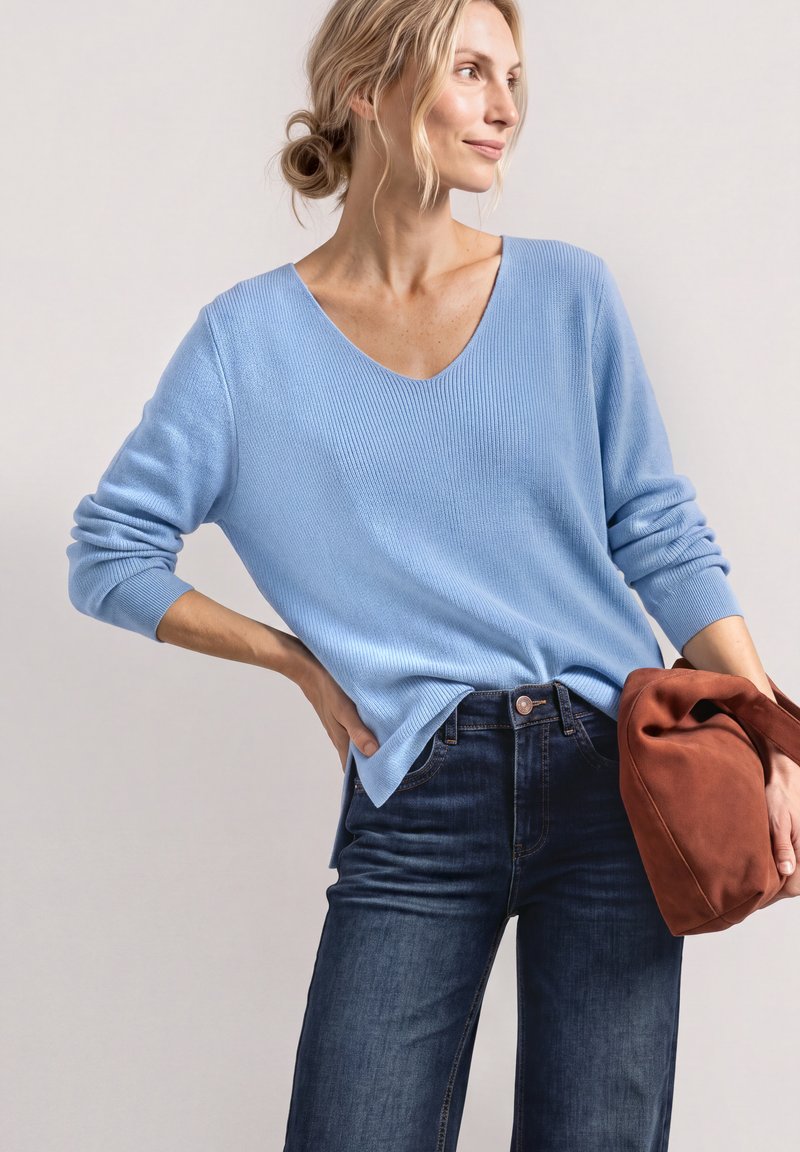 Femme aux cheveux blonds coiffés en chignon lâche, portant un pull bleu clair à col en V et un jean foncé, tenant un sac souple couleur rouille sur fond clair.