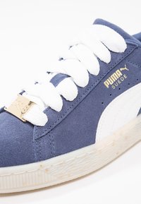 Gros plan sur une basket Puma en suède bleu avec des lacets blancs, une bande latérale blanche, un logo doré et une semelle blanche texturée parsemée de petites taches brunes.