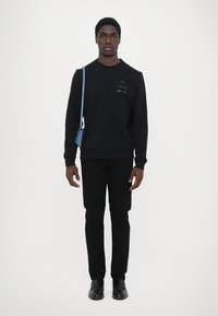 Sudadera negra con logo bordado colorido en el pecho, pantalones negros y zapatos negros. Bolso cruzado azul claro llevado sobre el hombro izquierdo.