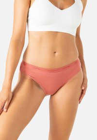 Femme portant un soutien-gorge de sport blanc et un slip de bain corail, se tenant contre un fond blanc.