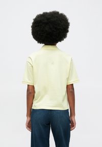 Άτομο με σγουρά μαλλιά που φοράει ένα ανοιχτό κίτρινο κοντομάνικο μπλουζάκι και μπλε τζιν, στέκεται με την πλάτη προς την κάμερα σε έναν απλό φόντο.