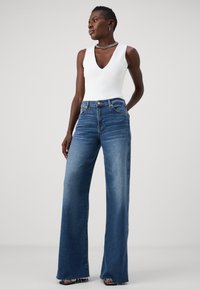 Jeans in denim blu a gamba larga con vita alta, dotati di un classico design a cinque tasche e una leggera scoloritura. Abbinati a un body bianco senza maniche.