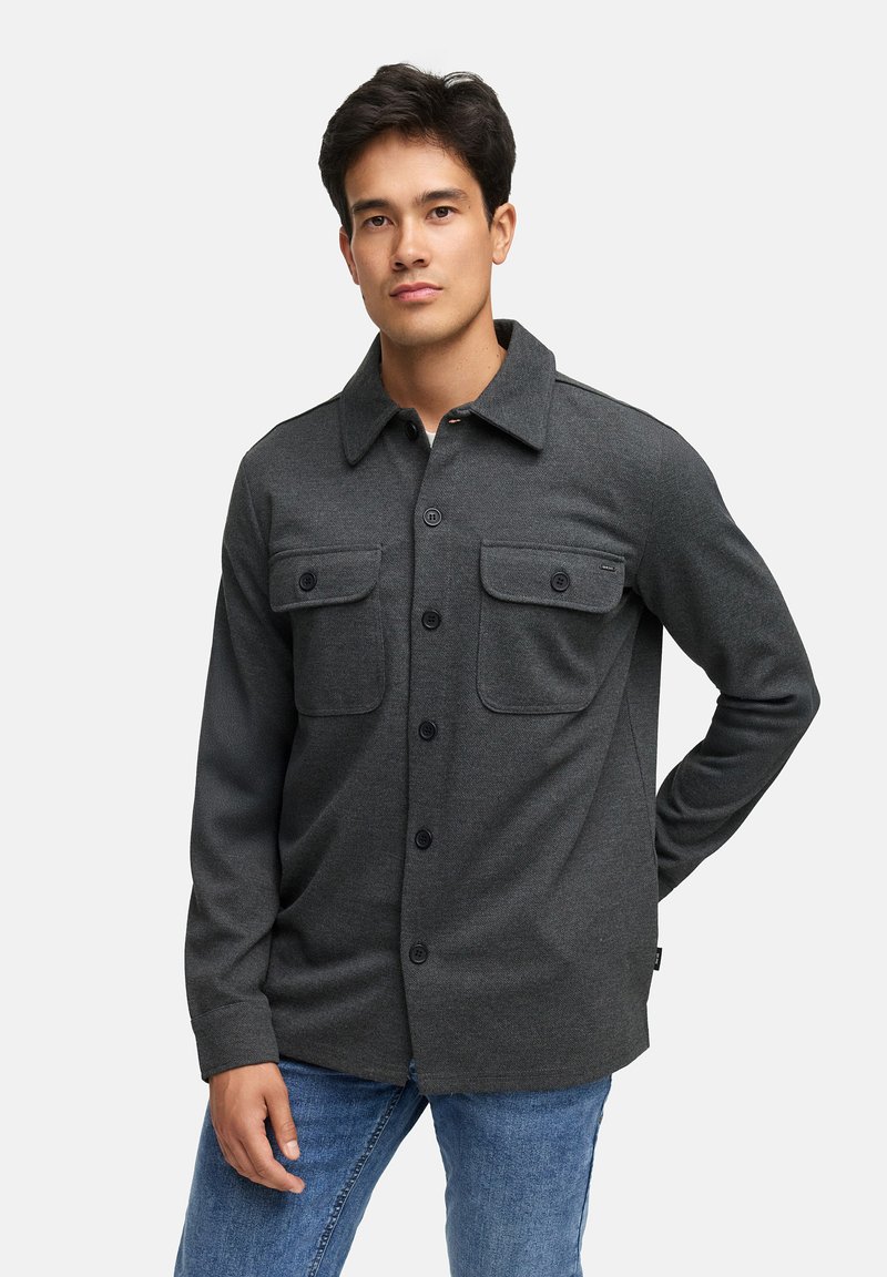 Grå skjorte med knapper, to brystlommer, lange ermer og spiss krage, laget av et strukturert stoff. Kombinert med blå jeans.