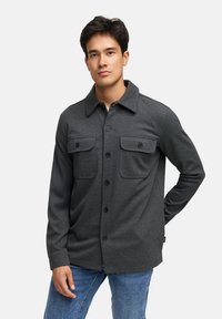 Grå skjorte med knapper, to brystlommer, lange ærmer og spidse kraver, lavet af et tekstureret stof. Bærer sammen med blå jeans.