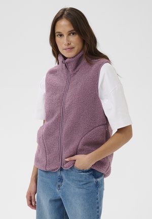 Donna che indossa un gilet in pile lavanda con zip sopra una maglietta bianca a maniche corte e jeans blu, in piedi con una mano in tasca.