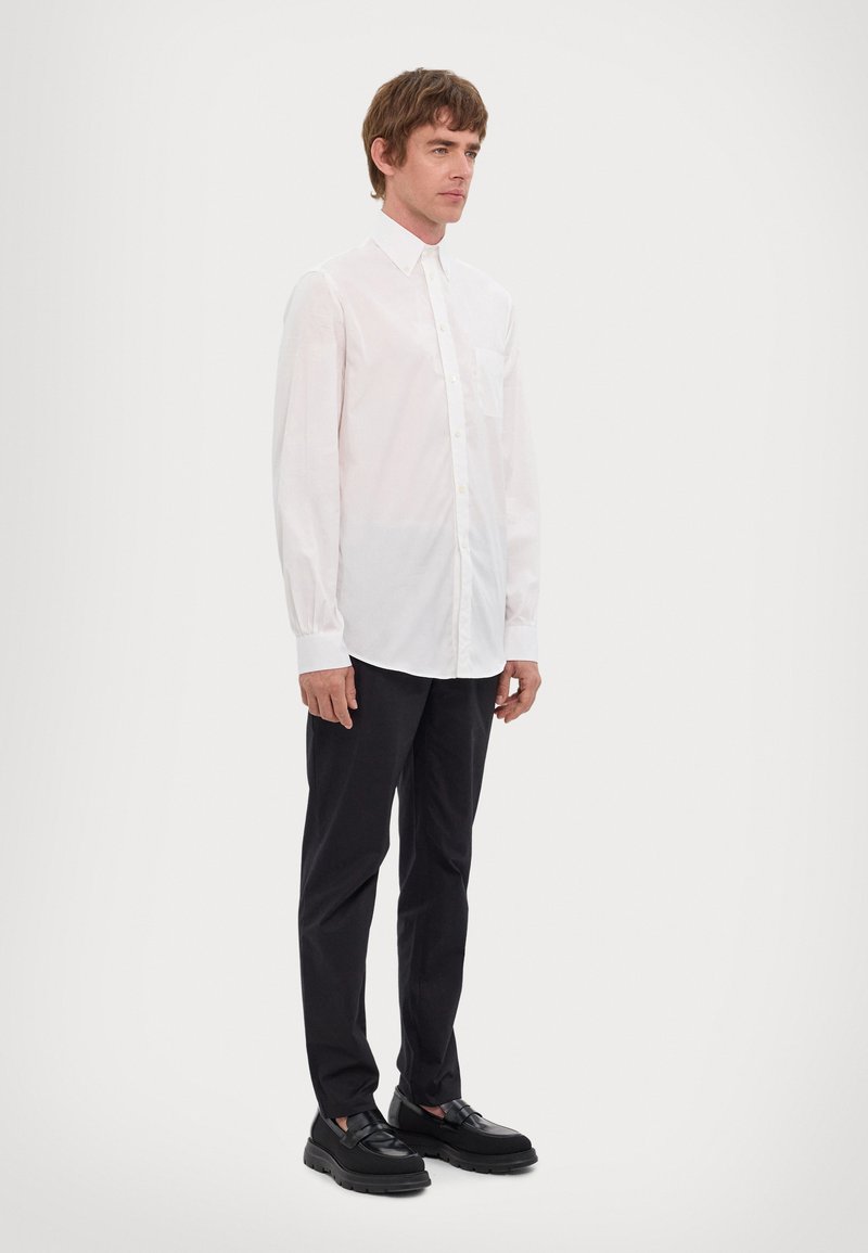 White long-sleeve shirt with button closure and pocket, paired with black trousers and black shoes featuring a thick sole.