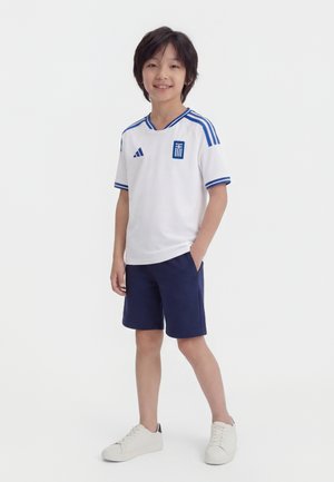 Ragazzo giovane in piedi, indossa una maglia sportiva bianca con strisce blu e pantaloncini blu scuro, sorride con una mano in tasca, scarpe da ginnastica bianche.