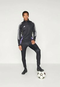 Svart idrettsdrakt med hvite og lilla striper, med glidelås krage. Modellen står på ett bein med en fotball ved føttene.