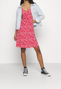 Czerwona sukienka w kwiaty z dekoltem w serek i marszczeniami, w zestawieniu z jasną dżinsową kurtką i czarnymi sneakersami za kostkę. Gładka tekstura materiału.