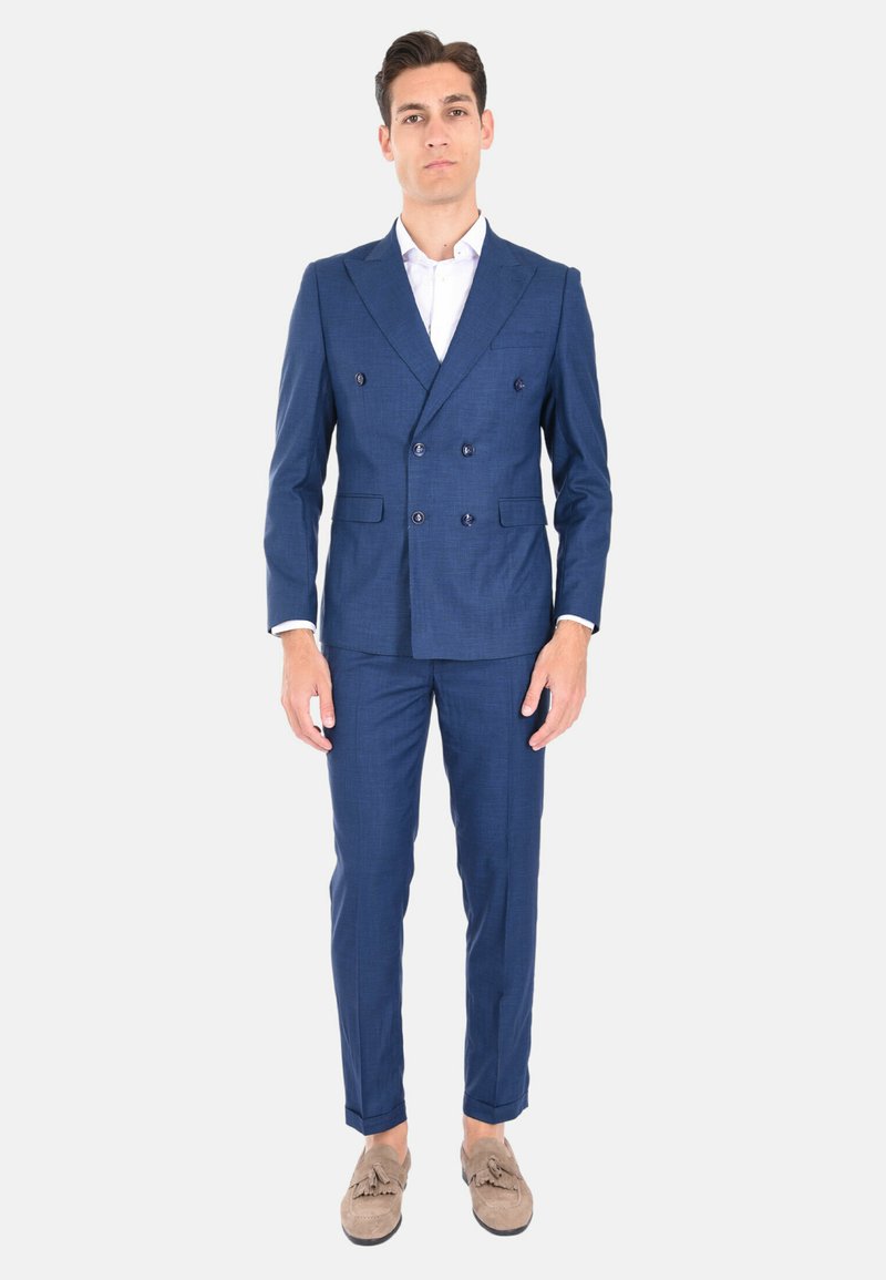 Giacca da abito doppiopetto in tessuto blu con rever a punta, abbinata a pantaloni slim-fit e mocassini beige con dettagli frangi.