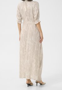 Femme portant une robe maxi beige à motif marbré ceinturée avec des manches 3/4 et des talons slingback beiges, se tenant devant un fond blanc uni.