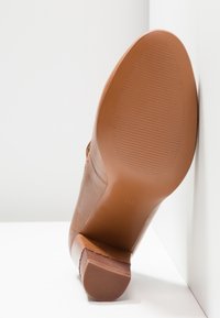 Talons en cuir marron avec un bout lisse et arrondi et une semelle en caoutchouc texturée. Le talon est de hauteur moyenne avec un design empilé.