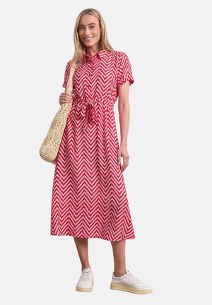 Mujer con vestido midi rojo y blanco con patrón de chevrones y mangas cortas, sujetando un bolso tejido beige y zapatillas blancas.