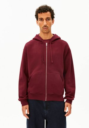 Mann trägt einen dunkelroten Kapuzenpullover mit Reißverschluss, Kordeln und Fronttaschen, kombiniert mit dunkelblauen Hosen, steht vor einem schlichten weißen Hintergrund.