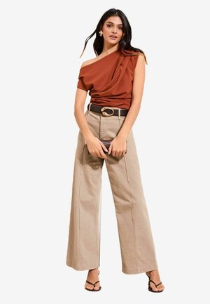 Kobieta ubrana w rdzawy top na jedno ramię, beżowe szerokie spodnie z czarnym paskiem, trzymająca małą ciemną kopertówkę, stojąca w czarnych sandałach.