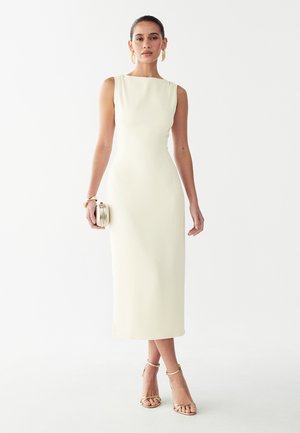 Mujer con vestido midi sin mangas color marfil, sujetando un pequeño bolso de mano dorado, con pendientes dorados, pulsera y sandalias de tacón alto con tiras.