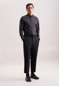 Camicia button-up grigia scura con colletto, abbinata a pantaloni neri a vita alta con pieghe e scarpe nere stringate, in piedi contro uno sfondo chiaro.