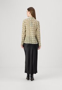 Blusa a maniche lunghe con stampa geometrica verde e oro, abbinata a una gonna nera in satin lunga fino al pavimento e stivaletti neri con tacco.