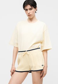Femme portant un t-shirt ample crème avec un short en tricot assorti orné de bordures noires et du logo Adidas, debout devant un fond uni.
