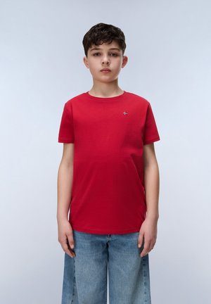 Boy standing facing forward, wearing a red short-sleeve t-shirt and light blue jeans against a plain light background.