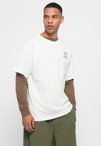 Hvid bomulds t-shirt med grøn og orange logo på venstre bryst, båret over en brun langærmet skjorte. Model i løse grønne shorts.