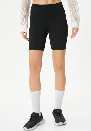 Shorts de vélo noirs en tissu extensible, dotés d'une taille haute et d'une texture lisse. À porter avec des chaussettes blanches jusqu'aux genoux et des baskets noires.
