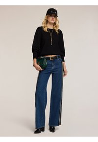 Maglione nero a coste, jeans in denim blu a vita alta con strisce laterali, stivali neri con tacco e una borsa verde. Cappello nero e gioielli dorati.