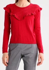 Rode trui met lange mouwen, met een gestructureerde gerimpelde kraag en geribbelde manchetten, gecombineerd met grijze, op maat gemaakte broek. Gladde stofafwerking.