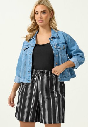 Woman wearing a light blue denim jacket over a black top and black-and-white vertical striped shorts, standing with one hand on her waist.