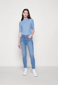 Camisola de manga curta azul claro emparelhada com jeans de cintura alta e corte slim, também em azul claro. A modelo está de pé com uma mão na cintura, usando ténis brancos.