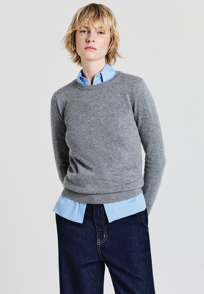 Pull en tricot gris à col rond porté par-dessus une chemise à col bleu clair. Un pantalon en denim foncé complète la tenue.