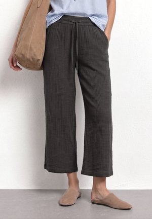 Personne portant un pantalon noir ample à cordon de serrage, une chemise rayée bleu clair, des chaussures beiges sans lacets, et portant un grand sac à main beige sur une épaule.